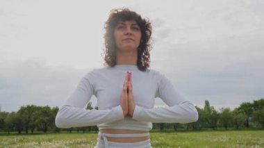 Aktif beyaz elbiseli kızın orta pozu parkta meditasyon yapmaya odaklanmış. Spor kadın avuçları birlikte yoga yapıyor. Sağlıklı yaşam tarzı kavramı. Asana pozisyonu. Yüksek kalite 4k görüntü