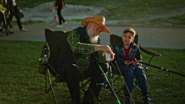祖父は孫に川沿いの釣り場にウジを置く方法を示した 小さな孫に魚を捕まえ 椅子に座ることを教える年金受給者男性 高品質の4K映像 — ストック動画