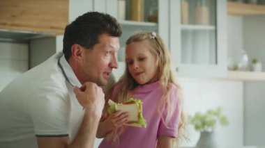 Neşeli, gülümseyen küçük bir kız mutfakta babasının yanında sandviç yiyor. Baba gülüyor ve kızına komik hikayeler ya da anekdotlar anlatıyor. İşaret parmağı hareketi gösteriliyor. Yüksek kalite 4k görüntü