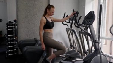 Mükemmel formda genç ve narin bir kadın spor salonuna girer ve bacak egzersizi makinesini özelleştirir. El hareketi ve pedal çevirme. Spor yaşam tarzı ve sağlıklı kavramı
