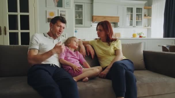 一个美国梦中的家庭坐在沙发上的房间里 父母为女儿选择未来的幼儿园或学校 妻子认真地听丈夫讲话 小女孩低着头点点头 — 图库视频影像