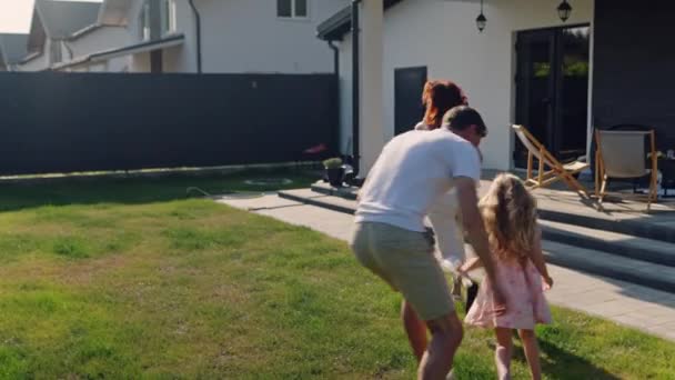 アメリカの夢の家族は家の近くの芝生の屋外で走り 晴れた夏の週末にリラックスします 父は娘を捕まえてスピンする 遊び心と健康的なアクティブライフスタイルのデモンストレーション イギリス — ストック動画