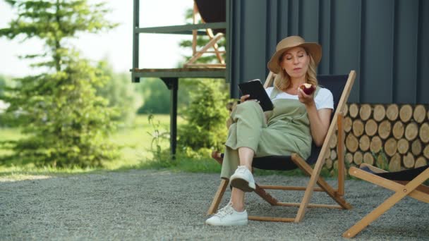 一位迷人的中年妇女在乡间别墅度过暑假 享受户外活动 下班后在花园休息 吃苹果 在平板电脑上读电子书 坐在躺椅上 — 图库视频影像