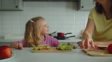 Mutlu anne, sandviçleri pişirme sürecini detaylıca anlatıyor. Kız bir gülümsemeyle dinler ve ona bakar. Mutlu çocukluk ve annelik kavramı. Kapatın. Yüksek kalite 4k