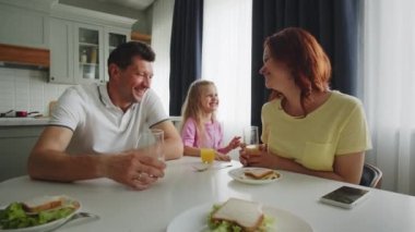 Mutfakta gülümseyen bir Amerikan rüya ailesi sandviç yer, meyve suyu içer, eğlenir, güler ve şakalar yapar. Kızı komik el hareketleri gösteriyor ve ebeveynleri eğlendiriyor. Mutlu çocukluk kavramı ve...