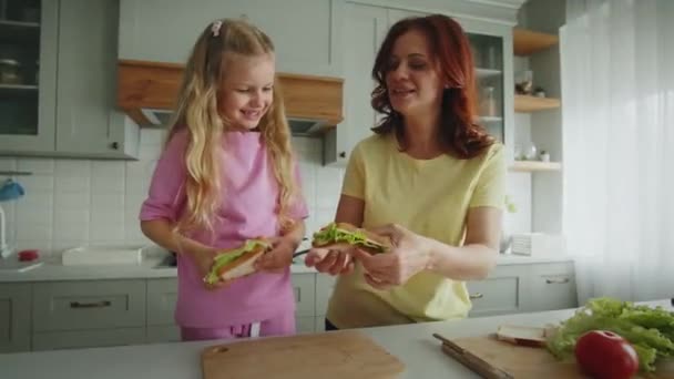 笑顔の母親と娘は キッチンで完成したサンドイッチを互いに示しています 暖かい家族の雰囲気の中でシンプルな料理の傑作のデモンストレーション 幸せのコンセプト — ストック動画