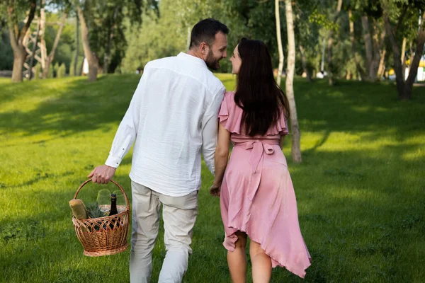 Rear view of young flirting couple walking and kissing on picnic. Happy couple holding hands together bringing basket fresh products. Family vacation outdoors. Cute pair resting in nature. High