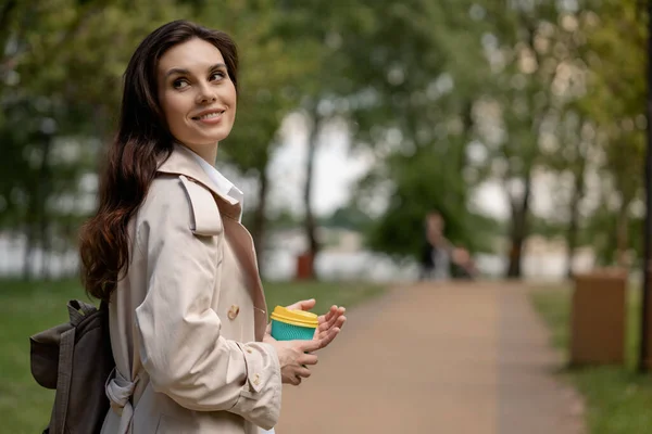 一位年轻漂亮的女士正在一个春天公园里散步 在外面喝杯咖啡 一个迷人的黑发女孩在一个熟人的召唤下转过身来 微笑着 一位女士对此感到高兴 — 图库照片