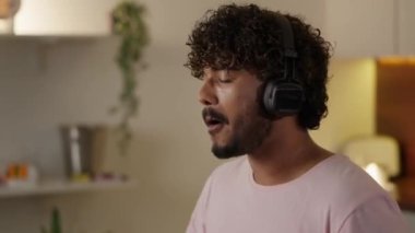 Sağlıksız beslenme kavramı. Müzik dinlerken fast food yiyen komik adam. Adam mutfakta lezzetli hamburger ve patates kızartmasının tadını çıkarıyor. Hintli erkek aldatma yemeklerine bayılır. Yüksek kalite 4k görüntü