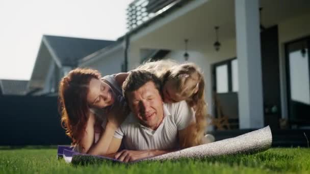 アメリカンドリームファミリーは 芝生の家の近くの屋外でリラックスし 紫色の毛布 カーペットや敷物の上に横たわります 父と母と娘はお互いに愛を示しています ママと子供はキスして パパを抱きしめる イギリス — ストック動画