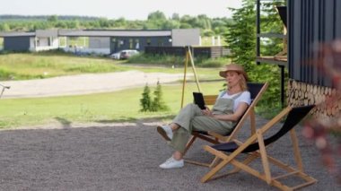 Orta yaşlı bir kadın yaz tatilini kır evinde geçirir, açık havadan ve işten sonra bahçede dinlenerek, sakin bir ortamda bir koltukta oturur. Bir bayan okumak için tablet kullanıyor.