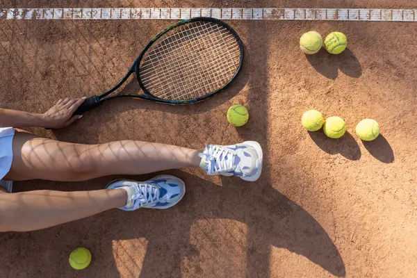 Top view of black tennis racket, balls and sports woman legs in trainers. Sport equipment lying on tennis court. Sports woman wear mini skirt holding racquet near balls. High quality photo