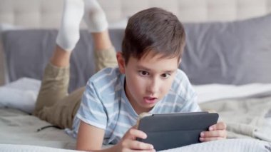 Küçük bir çocuk yatak odasındaki kanepede karnının üzerine yatar ve bir tablet kullanır. Bir çocuk online oyun oynuyor özenle ve duygusal bir şekilde kızgın ve alaycı bir gülümseme sergiliyor, kaybettiği için aleti sallıyor.