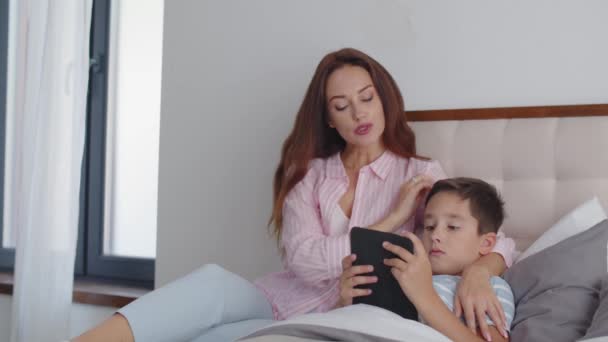 男の子はベッドの上に横たわり 寝る準備をし タブレットの電子ブックを読みます 若くて思いやりのある母親が息子の隣に座り 意識を持って頭をストロークします 家族における反応の概念 — ストック動画