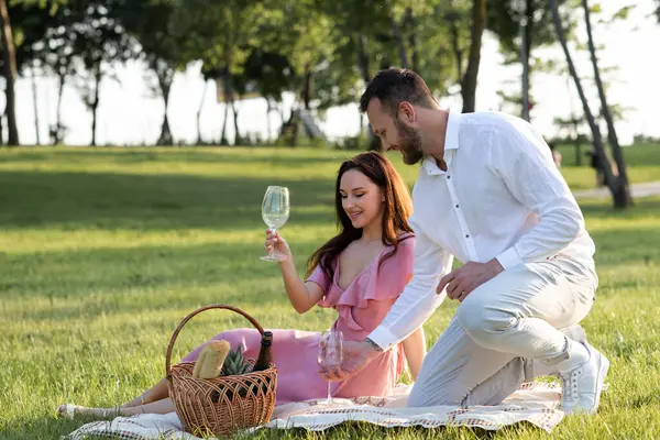 Man preparing romantic place for a date with beautiful girlfriend. Boyfriend serving wine glasses. Romance picnic in park. Happy family lunch outdoors. Lovely couple sitting at the blanket outside