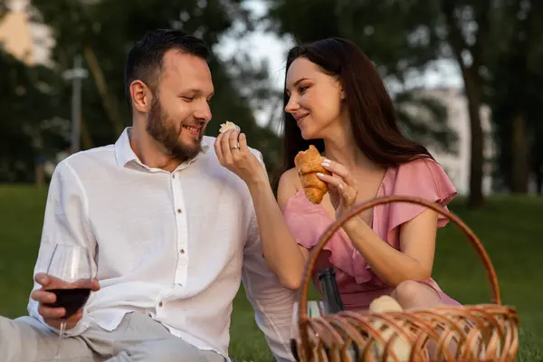 Cute young girlfriend feeds her boyfriend french yummy croissant on picnic. Couple having romantic date outdoors. Wife giving husband a small piece of bakery in park. Family vacation. High quality