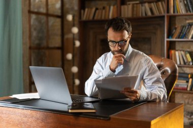 Ciddi bakışları olan ve önemli belgeleri dikkatle okuyan bir adam kütüphanede bir masaya oturur ve dizüstü bilgisayar kullanır. Saygıdeğer bir iş adamı karlı bir kontrata aşina. Bir genç.