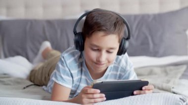 Küçük bir çocuk yatak odasındaki kanepede yüzüstü yatıyor, tablet ve kulaklık kullanıyor. Bir çocuk online oyun oynuyor özenle ve duygusal olarak, kızgın olduğunu gösteriyor ve yüzünü buruşturuyor, aleti sallıyor.
