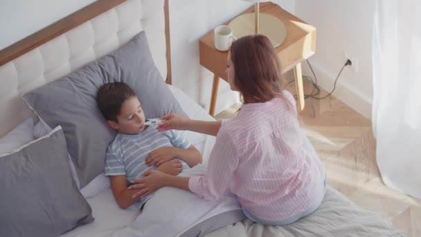 病童感觉不适 测量体温 躺在卧室里 保持卧床休息 一位有爱心的年轻母亲坐在旁边 给儿子数字温度计 摸他的额头检查 — 图库视频影像