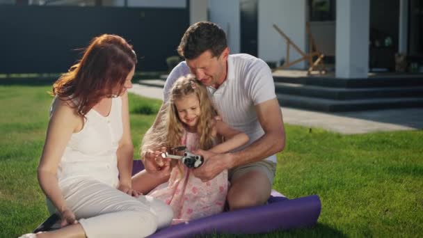 ジョイフルなアメリカンドリームファミリーは 紫色の毛布 カーペットや敷物の上の家の近くの芝生の屋外に座っています 親と娘は笑顔 カスタマイズ ショーで玩具ドローンを起動しています — ストック動画