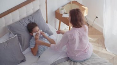 Hasta bir çocuk öksürüyor ve ağızını eliyle kapatıyor, burnunu tek kullanımlık bir mendile siliyor, yatak odasına uzanıyor ve yatak istirahatı sağlıyor. Şefkatli genç bir anne yanına oturur, peçete verir ve oğlunun alnına dokunur.