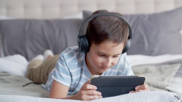小さな男の子が寝室のソファーに横たわり タブレットとヘッドフォンを使用しています 子供は注意深くオンラインゲームをプレイし グリムを作り わずかな皮肉な笑顔を示し デバイスを揺さぶります — ストック動画