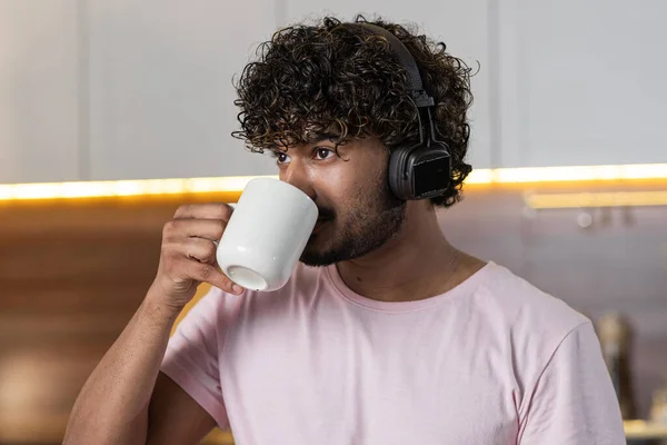 A mestizo guy stands sideways, holds a cup of tea or coffee in hand and drinks it, listens to music on headphones and looks into the distance. An american or african young man enjoys a hot beverage in
