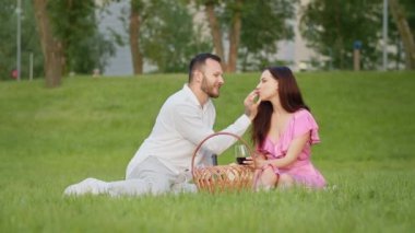 Battaniyenin üzerinde oturmuş dışarıda romantik bir piknik yapan neşeli elbiseli genç bir aile. Koca, karısını kırmızı şarap içerken parkta besler. İlahi ilişki yıldönümü kavramı. Yüksek