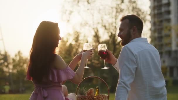 若い家族カップルは 都市公園で夕日を見ているロマンチックなピクニックディナーを持っています 赤ワインを飲む男性と女性がアウトドアで最初の記念日を祝います 地元の芝生の日付 高品質4Kについて — ストック動画