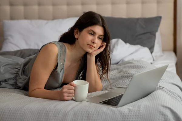 ベッドの上に横たわり 朝寝室でラップトップと仕事をしているドリーミーなブルネットの少女 高品質の写真 — ストック写真