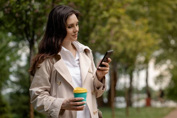 若い女性は 指定された時間に友人や職場の同僚に会うのを待って スマートフォンを使用したり メッセージを読んだりします ブルネットの女性がスプリングパークでコーヒーを飲みながら散歩 — ストック写真