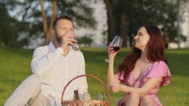 Güzel beyaz bir çift piknikte şarap içer. Açık havada rahatlamış bir aile. Parkta birlikte dinlenen beyaz şarap kadehlerini tutarak. Kadın ve erkek şenlikli kıyafetler giyer ve battaniyeye otururlar.