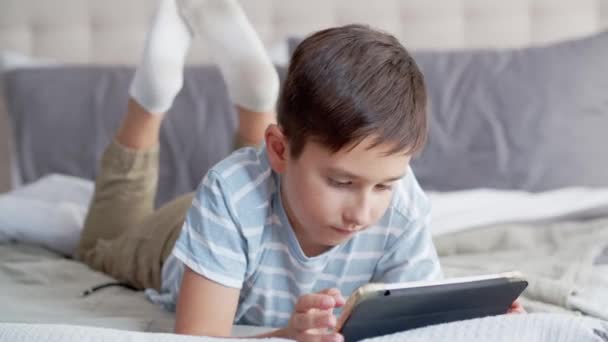 小さな男の子は寝室のソファーの上に横たわり タブレットを使用しています 子供が熱心かつ感情的にオンラインゲームをプレイし グリムを作り 攻撃を示し 揺さぶります — ストック動画