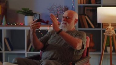 Sağlıklı ve pozitif ruh hali olan yaşlı bir adam bir koltukta oturur, akıllı telefon ve görüntülü konuşma kullanır. Bir insan dalgası selamlama işareti olarak el sallar, gülümser, hareketli konuşur ve...