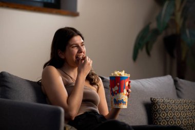 Heyecanlı genç kadın televizyon izlerken kanepede patlamış mısır yiyor. Kapalı mekânda çekilen kız abur cuburlardan hoşlanır. Yüksek kalite fotoğraf