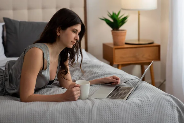 美しいフリーランサーの少女がベッドに横たわってコーヒーを飲み 朝はラップトップで仕事をする 高品質の写真 — ストック写真