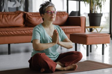 Orta yaşlı bir bayan evde yoga yapıyor. Gözleri ve avuçları kapalı bir kadın, göğsünde katlanarak, lotus pozisyonunda yerde bir paspasın üzerine oturur. Manevi gelişim kavramı