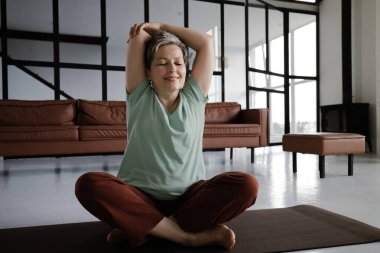 Orta yaşlı bir kadın evde yoga yapıyor. Mutluluktan gözleri kapalı gülümseyen bir kadın lotus pozisyonunda yerde bir paspasın üzerine oturur ve kollarını başının üstüne uzatır. Vücudun uyumu kavramı