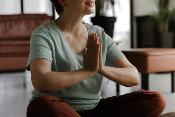 A middle aged lady is practicing yoga at home. A woman without a face sideways sits on the floor in the lotus position with palms, folded together on chest and meditates. Close up. Selective focus