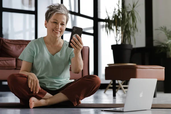 A middle aged lady rests after training yoga in the large hall. A happy woman sits on a mat on the floor, uses a smartphone and laptop, has video call with a meditation instructor or takes a selfie