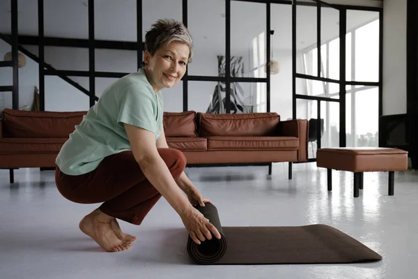 A middle aged lady practices yoga at home. A smiling woman in a satisfied mood squats sideways and barefoot on tiptoes, rolls up the mat after training. Close up. Looks at the camera. High quality