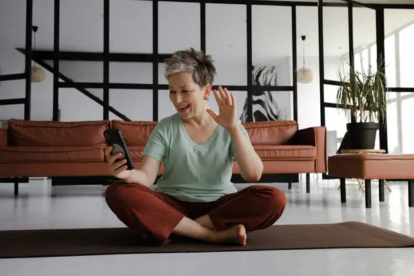 A middle aged lady practices yoga in the large hall of the house. A smiling woman sits on a mat on the floor in the lotus position, has a break time from training, uses a smartphone for video