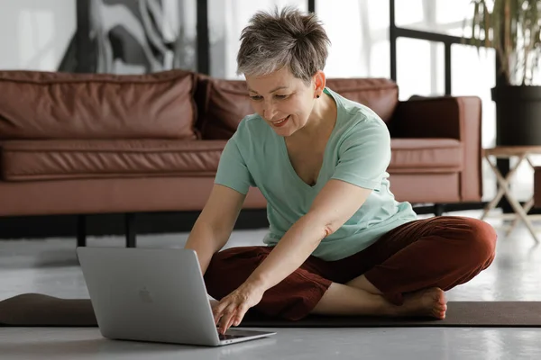 A middle aged lady is practicing yoga at home. A smiling woman sits on a mat on the floor in the lotus position, has a break time from training, uses a laptop and laughs, watching funny distracting