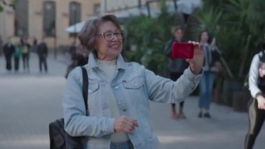 Neşeli bir ruh hali içinde gülümseyen yaşlı bir kadın sıcak bir bahar gününde turistik bir şehirde kaldırımda dikiliyor. Zarif bir bayan akıllı telefon kullanır, görüntülü konuşma yapar, araziyi ve onu gösterir.