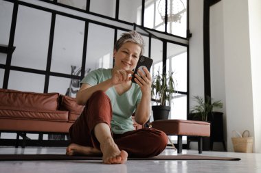 Orta yaşlı bir bayan, evin büyük salonunda yoga eğitimi aldıktan sonra dinleniyor. Gülümseyen bir kadın yerde bir paspasın üzerinde oturur, iletişim kurmak için akıllı telefon kullanır, ilginç şeyler arar ve okur.