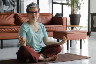 Orta yaşlı bir bayan evde yoga yapıyor. Yüzünde hafif bir gülümseme olan bir kadın lotus pozisyonunda yerde bir paspasın üzerine oturur. Fiziksel egzersiz yoluyla ruhsal gelişim kavramı