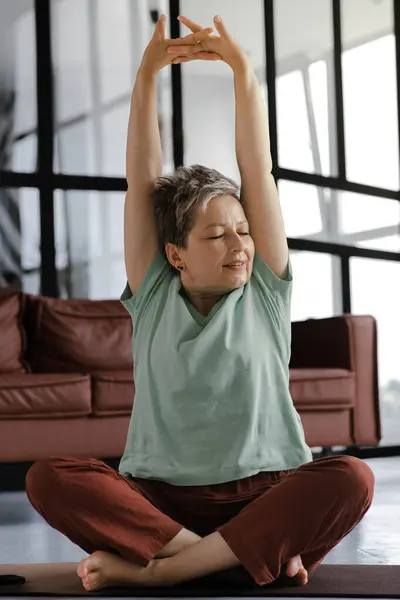 A middle aged lady practices yoga at home. A woman with closed eyes in bliss sits on a mat on the floor in the lotus position, stretches arms above head with palms, facing the ceiling, and fingers