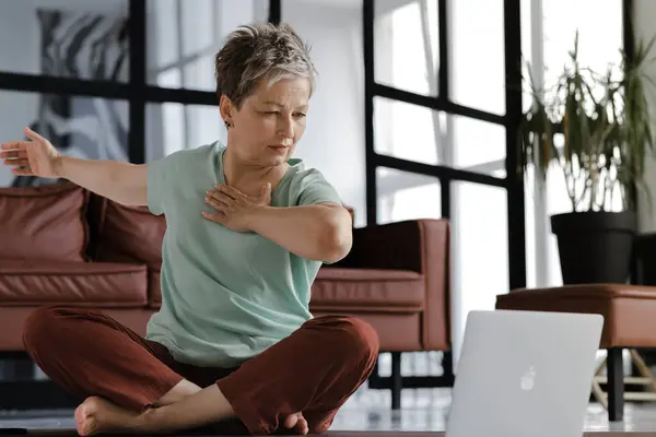一位中年妇女在家里练习瑜伽 一位长相凝神的妇女坐在地板上的一块垫子上 她的身体保持着莲花的姿势 她把胳膊挪到旁边 用笔记本电脑 看着练习 然后重复练习 — 图库照片