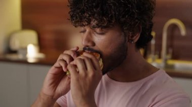 Sağlıksız yemek teslimatı kavramı. Hintli adam hamburgerinin tadını çıkarıyor. Sapık saçlı genç adam mutfakta lezzetli bir burger yiyor. Erkek öğrenci büyük bir sandviç yiyor. Yüksek kalite 4k görüntü