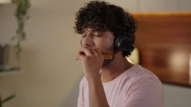 Sağlıksız beslenme kavramı. Müzik dinlerken fast food yiyen komik adam. Adam mutfakta lezzetli hamburger ve patates kızartmasının tadını çıkarıyor. Hintli erkek aldatma yemeklerine bayılır. Yüksek kalite 4k görüntü